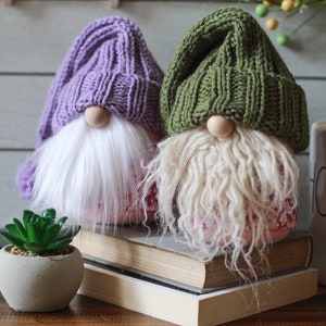 May include: Two knitted gnomes with white beards and pink floral patterned bases. The gnome on the left is wearing a purple hat and the gnome on the right is wearing a green hat. The gnomes are sitting on a stack of books.
