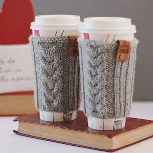 May include: Two grey knitted coffee cup sleeves with brown leather labels. The labels read "Good Vibes Only" and "Coffee Queen". The sleeves are on top of a brown book.