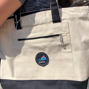 May include: A white canvas tote bag with black straps and a black bottom. The bag has a zippered pocket on the front and a round patch with a mountain scene and the text "SWA" in black.
