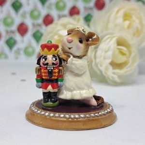 May include: A whimsical figurine featuring a mouse in a white dress and a nutcracker soldier. The mouse is standing next to the nutcracker on a round base with a decorative border. The figurine is a charming holiday decoration.