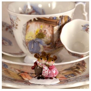 May include: Two small brown mice figurines wearing scarves and holding hands. The mice are standing on a white base with a pink heart design. The figurines are in front of a teacup and saucer.