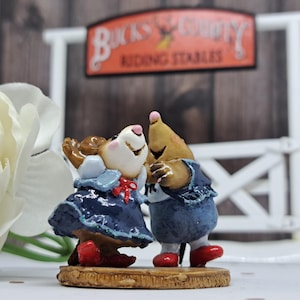 May include: A figurine of two cartoon mice in a loving embrace. The mouse on the left wears a blue dress with a red bow and red shoes. The mouse on the right wears a blue denim outfit and red shoes. The figurine is on a wooden base.