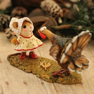 May include: A small, white-dressed mouse figurine with a red bow and a yellow flower in her hair looks up at a brown and white turkey figurine with a red wattle.