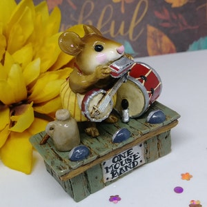 May include: A small, brown mouse figurine playing a banjo and a drum. The mouse is wearing yellow and white striped pants and is sitting on a wooden stage with a sign that reads "One Mouse Band".