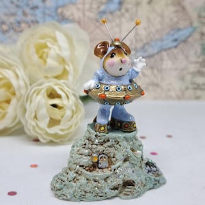 May include: A whimsical figurine of a mouse dressed in a blue and gold space suit, standing atop a glittery, textured rock formation. The mouse has large ears and is wearing a helmet with antennae. The figurine is a decorative item.