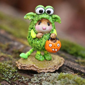 May include: A whimsical Halloween figurine of a mouse dressed in a green monster costume, holding an orange jack-o'-lantern treat bag. The mouse has large, googly eyes and is standing on a small, textured base. The figurine is set against a blurred, natural background.