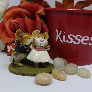 May include: A figurine of two mice, one in a tuxedo holding a heart-shaped box, kissing the other mouse in a white dress. A red pot with the word "Kisses" is in the background, along with decorative stones and a rose.