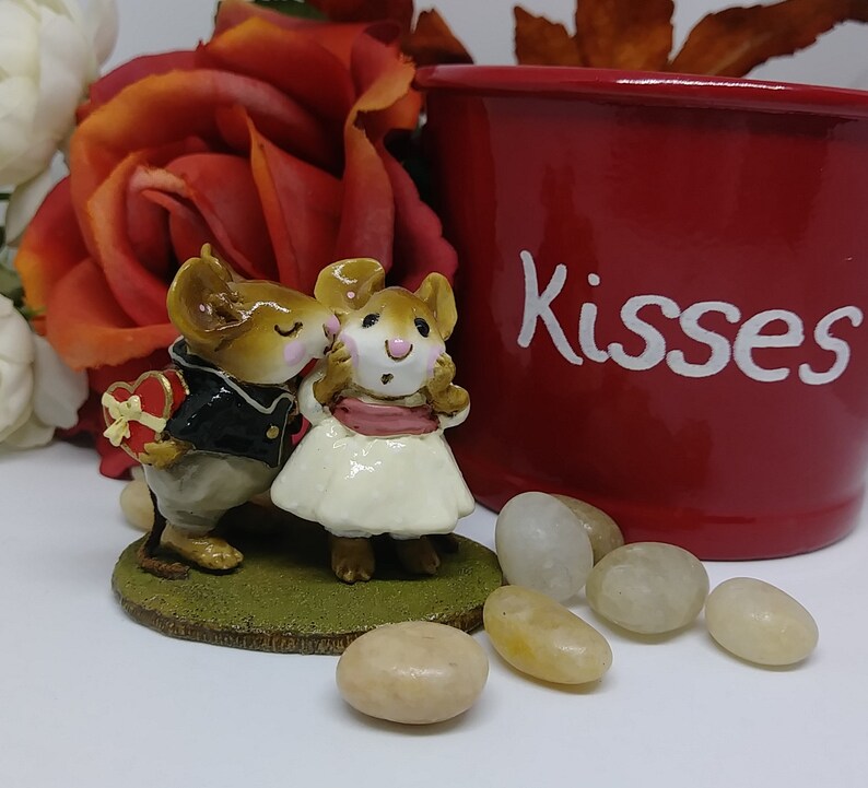 May include: A figurine of two mice, one in a tuxedo holding a heart-shaped box, kissing the other mouse in a white dress. A red pot with the word "Kisses" is in the background, along with decorative stones and a rose.
