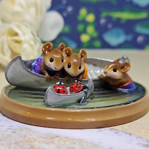 May include: A decorative figurine featuring three brown mice in a canoe, with two ladybugs in a smaller boat. The mice wear blue and orange clothing. The base is a light brown with a green water effect.