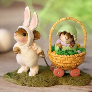 May include: A white bunny rabbit figurine wearing a white bunny costume pulls a basket with a mouse figurine inside an eggshell. The basket has wheels and is decorated with green grass and yellow eggs.