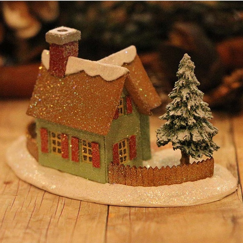 May include: A miniature Christmas house with a green exterior, red shutters, and a brown roof dusted with glitter. A small Christmas tree and a fence are also included in the scene. The house sits on a white, glittery base.