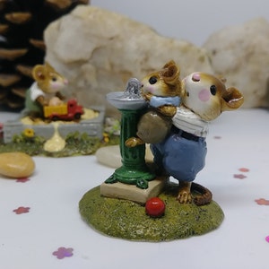 May include: Two brown mice figurines, one carrying the other, standing by a green water fountain. The mice are painted with details, including blue clothing and pink cheeks. The fountain is on a green base with a brown and green platform.