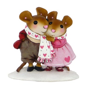 May include: A figurine of two brown mice wearing winter clothing. The male mouse is wearing a brown jacket, a red scarf, and a white scarf with pink hearts. The female mouse is wearing a pink dress and a pink scarf with white polka dots. The mice are standing on a white base.