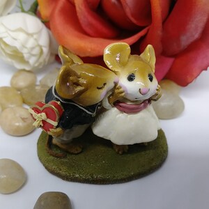 May include: A figurine of two mice in love. One mouse, wearing a black suit, holds a heart-shaped box. The other mouse, in a white dress, is being kissed. The figurine sits on a green base, with a red rose in the background.