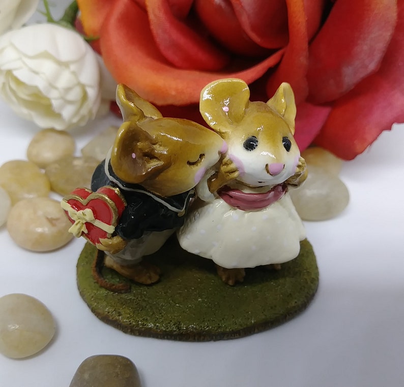 May include: A figurine of two mice in love. One mouse, wearing a black suit, holds a heart-shaped box. The other mouse, in a white dress, is being kissed. The figurine sits on a green base, with a red rose in the background.