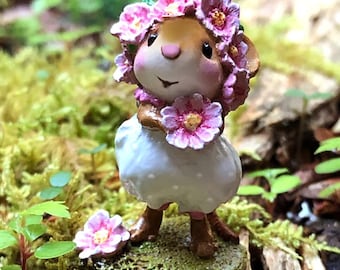 以下が含まれることがあります： 花冠をかぶり、花を持っているネズミの気まぐれな置物。 ネズミは白いドレスと茶色のブーツを着て、苔むした台座の上に立っています。 この置物は陽気な表情をしており、緑に囲まれています。