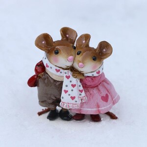 May include: A pair of brown and white mouse figurines wearing scarves and holding each other. The female mouse is wearing a pink dress with a heart design. The male mouse is wearing a brown jacket and a white scarf with pink hearts.