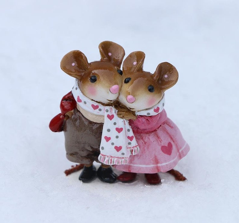 May include: A pair of brown and white mouse figurines wearing scarves and holding each other. The female mouse is wearing a pink dress with a heart design. The male mouse is wearing a brown jacket and a white scarf with pink hearts.
