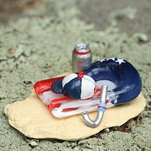 May include: A miniature scene of a red, white, and blue inflatable pool float with a blue and white baseball cap on top. A silver can and a blue and white snorkel are also included in the scene.