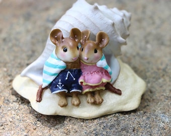 May include: Two small mouse figurines wearing beach clothes and a seashell, sitting on a sandy base.