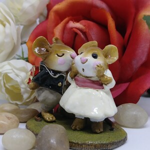 May include: A figurine of two mice, one in a black jacket and the other in a white dress, standing on a patch of green grass. The male mouse holds a bouquet. A red rose and white flowers are in the background.