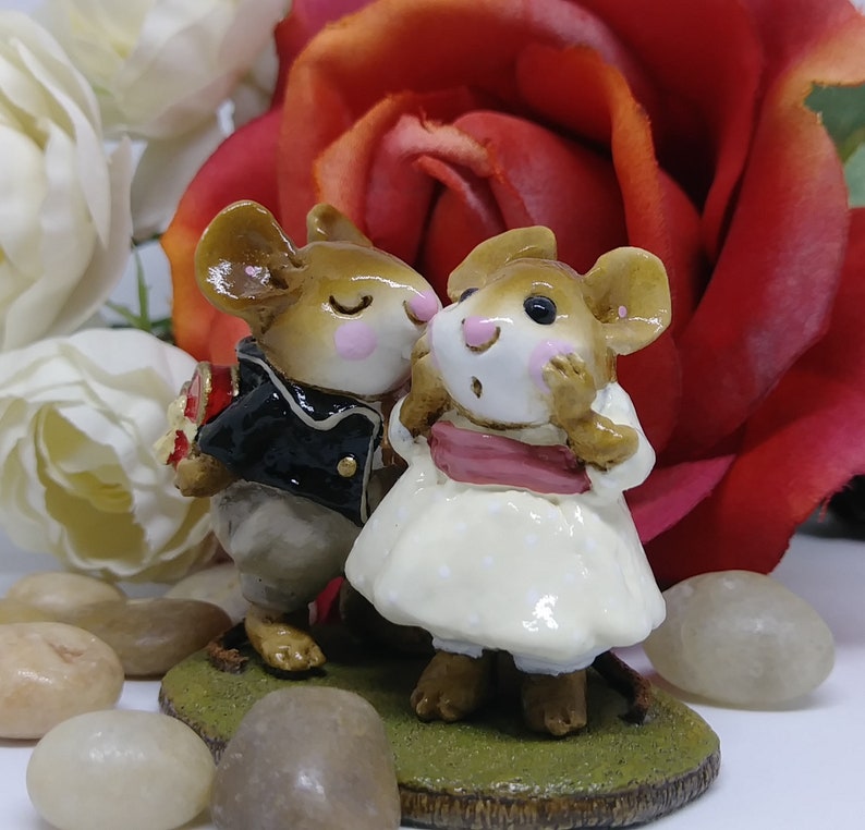 May include: A figurine of two mice, one in a black jacket and the other in a white dress, standing on a patch of green grass. The male mouse holds a bouquet. A red rose and white flowers are in the background.
