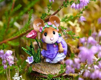 May include: A small, brown mouse figurine wearing a purple dress and a flower crown. The mouse is sitting on a small, brown mushroom-shaped base. The background is a blur of green leaves and purple flowers.