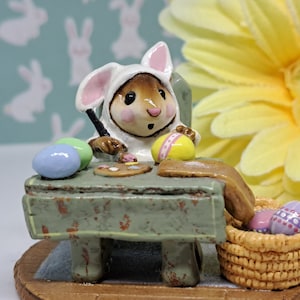 May include: A small bunny figurine wearing a white bunny costume is painting Easter eggs at a table. The bunny is holding a paintbrush and there are several colorful eggs on the table. There is a basket of eggs next to the table.