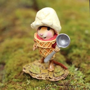 May include: A small figurine of a mouse wearing a waffle cone as a body and a scoop of white ice cream as a hat. The mouse has pink cheeks and holds a silver-colored metal spoon in its hand. The figure stands on a textured, leaf-shaped base.