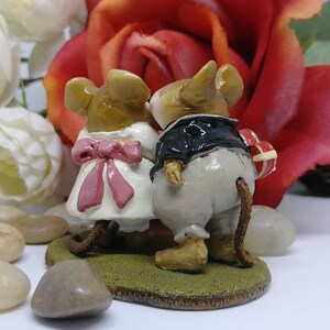 May include: A figurine of two mice, one in a white dress with a pink bow and the other in a black tuxedo, standing on a green base. The mouse in the tuxedo holds a heart-shaped gift. A red rose and white flowers are in the background.