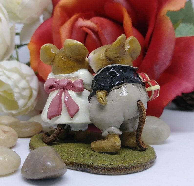 May include: A figurine of two mice, one in a white dress with a pink bow and the other in a black tuxedo, standing on a green base. The mouse in the tuxedo holds a heart-shaped gift. A red rose and white flowers are in the background.