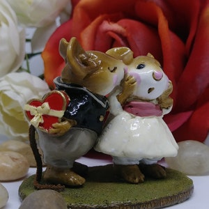 May include: A figurine of two mice in a romantic embrace. One mouse, dressed in a black jacket, holds a heart-shaped box. The other mouse wears a white dress with a pink sash. The figurine is set on a green base, with a red rose in the background.