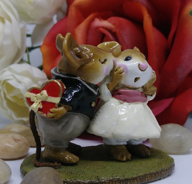 May include: A figurine of two mice in a romantic embrace. One mouse, dressed in a black jacket, holds a heart-shaped box. The other mouse wears a white dress with a pink sash. The figurine is set on a green base, with a red rose in the background.