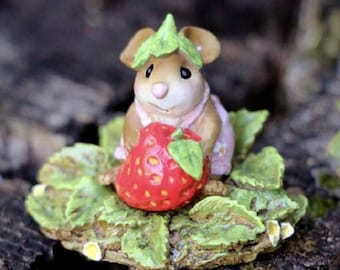 May include: A whimsical figurine of a mouse wearing a green leaf hat and a pink dress, holding a large red strawberry. The mouse is sitting on a bed of green leaves, creating a charming miniature scene.