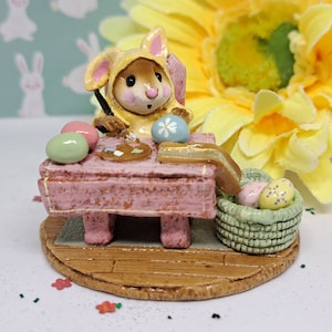 May include: A whimsical Easter figurine depicts a mouse in a bunny suit painting eggs. The mouse sits at a pink table with a palette and brush. A basket of decorated eggs and a large yellow flower complete the scene. The figurine is set on a wooden base.