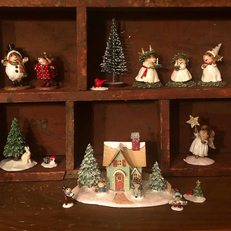 May include: A collection of Christmas figurines displayed in a wooden shelf. The figurines include snowmen, mice dressed in festive attire, a house, trees, and a red bird. The color palette is dominated by white, red, green, and gold.