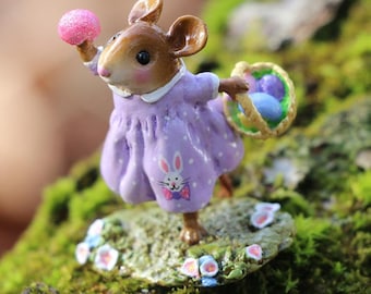 May include: A small, brown mouse figurine wearing a purple dress with white polka dots and a bunny rabbit design. The mouse is holding a pink egg in one hand and a basket of blue eggs in the other.