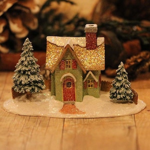 May include: A miniature Christmas village scene featuring a green house with a glittery gold roof and red door. Two small, snow-covered evergreen trees flank the house. The scene is set on a snowy base, evoking a festive holiday atmosphere.