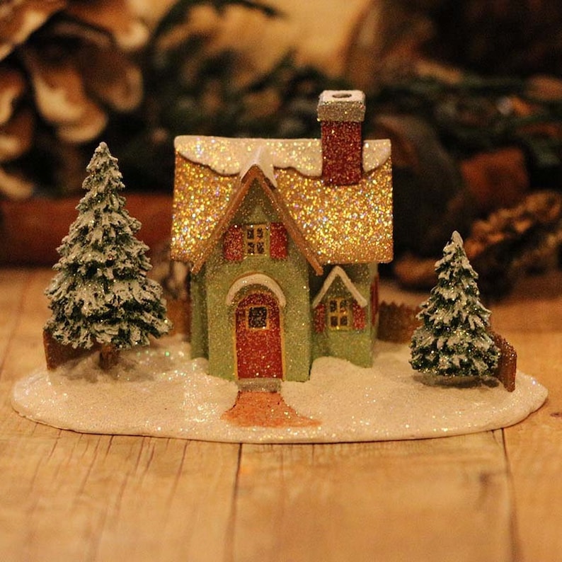 May include: A miniature Christmas village scene featuring a green house with a glittery gold roof and red door. Two small, snow-covered evergreen trees flank the house. The scene is set on a snowy base, evoking a festive holiday atmosphere.