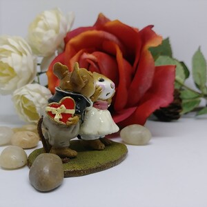 May include: A figurine of two mice in formal attire, one holding a heart-shaped box. The mice are kissing, set against a backdrop of a red rose and white flowers. The figurine is on a green base.