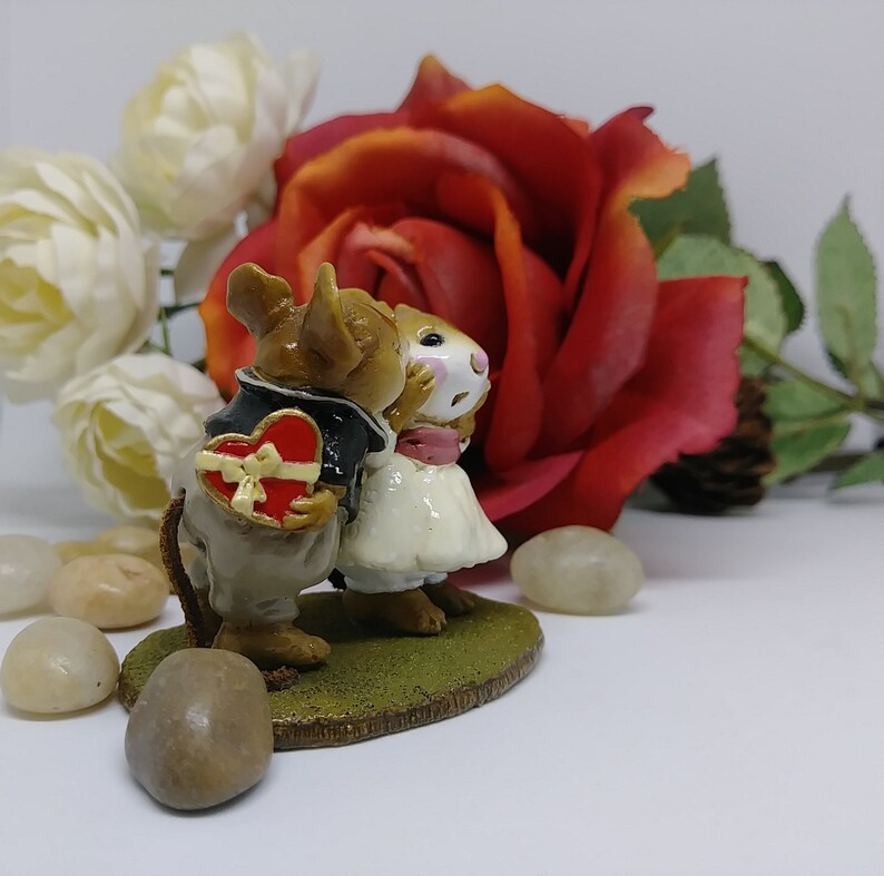 May include: A figurine of two mice in formal attire, one holding a heart-shaped box. The mice are kissing, set against a backdrop of a red rose and white flowers. The figurine is on a green base.
