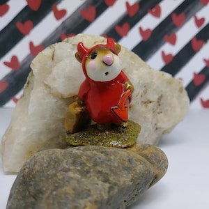 May include: A small figurine of a mouse dressed in a red devil costume with horns, holding a brown bag and a red arrow sign. The mouse stands on a patch of green grass atop a rock. The background features a black and white striped pattern with red hearts.