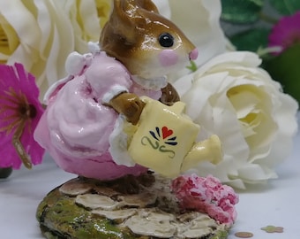 以下が含まれることがあります： ピンクのドレスを着て、赤いハートと青い花のデザインが施された黄色のジョウロを持った、小さな茶色のネズミの置物。ネズミは緑と茶色の土台の上に立っており、石の道があります。