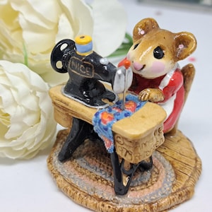 May include: A figurine of a mouse dressed in a red and white outfit, seated at a miniature sewing machine. The black machine has the word "NICE" on it. The mouse is sewing a blue and orange patterned fabric. The figurine is on a small rug.