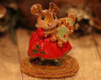 May include: A small, brown mouse figurine wearing a red dress with holly accents, holding a gingerbread person figurine. The mouse is standing on a woven straw base.