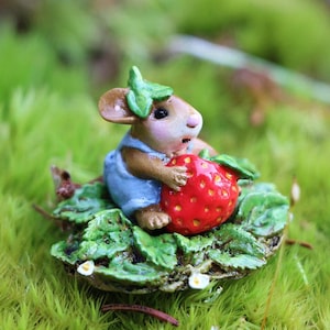 May include: A miniature figurine of a mouse wearing blue overalls, holding a large red strawberry. The mouse is sitting on a bed of green leaves and moss. The figurine is detailed and whimsical.