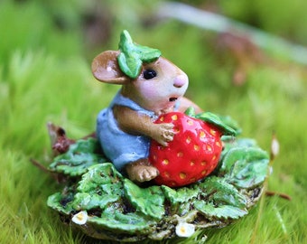 May include: A miniature figurine of a mouse wearing blue overalls, holding a large red strawberry. The mouse is sitting on a bed of green leaves and moss. The figurine is detailed and whimsical.