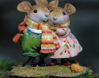 May include: A figurine of two mice wearing winter clothing. The mouse on the left is wearing a green and brown outfit with a yellow and orange scarf. The mouse on the right is wearing a pink dress with a red berry pattern and a white scarf. The mice are standing on a mossy base.