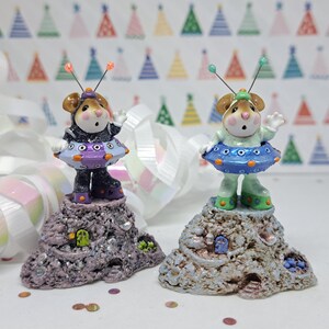May include: Two small, hand-painted figurines of mice dressed as astronauts. The mice are standing on small, rocky bases with a sparkly finish. The mouse on the left is wearing a purple and orange spacesuit with a black and white pattern. The mouse on the right is wearing a blue and green spacesuit with a white and orange pattern.