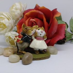 May include: A figurine of two mice in formal attire, one holding a heart-shaped box. The female mouse wears a white dress and pink sash. A large red rose and white roses are in the background, with small stones in the foreground.